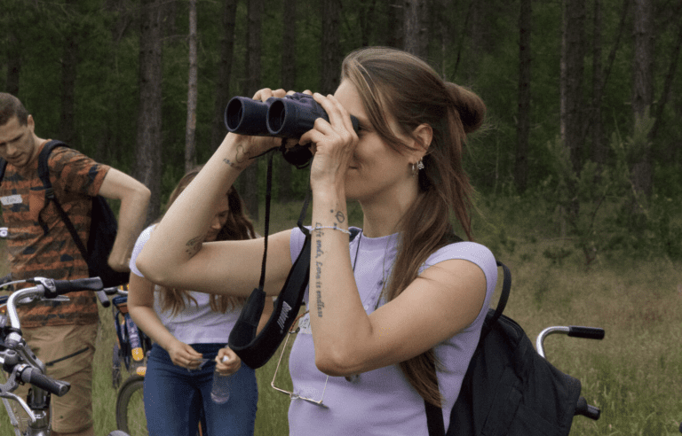 Een vrouw kijkt door een verrekijker in een bos; fietsen en mensen op de achtergrond.