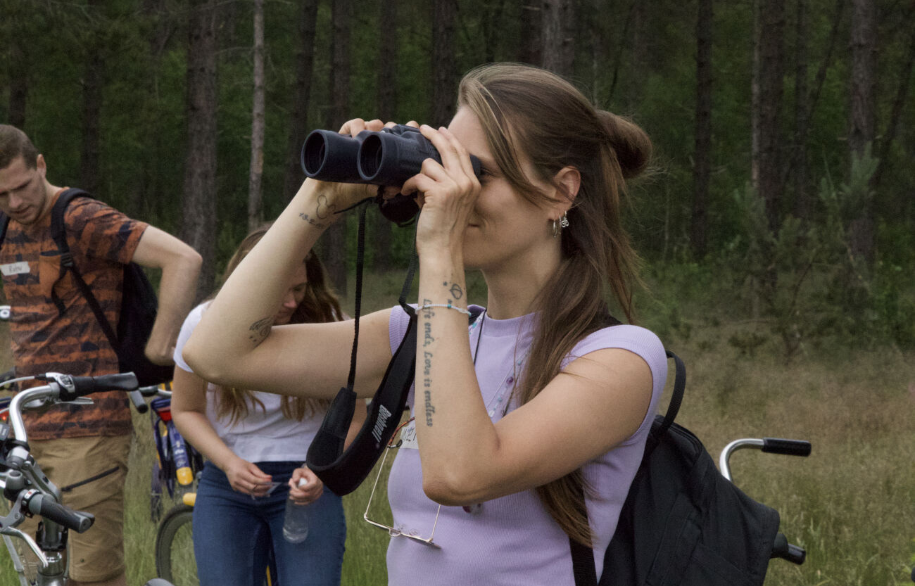 Een vrouw kijkt door een verrekijker in een bos; fietsen en mensen op de achtergrond.