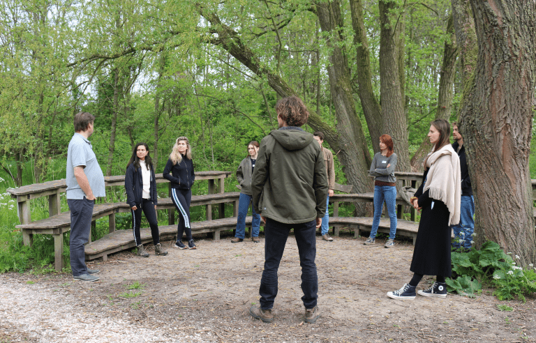 Groep mensen in een kring in een bosrijke omgeving, pratend en omgeven door bomen.