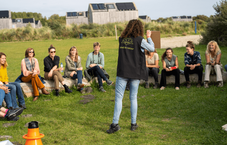 Persoon leidt buiten een groep mensen die op boomstammen zitten, met houten gebouwen op achtergrond.