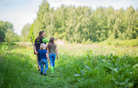Man en twee kinderen wandelen in een groen veld, bomen op de achtergrond.