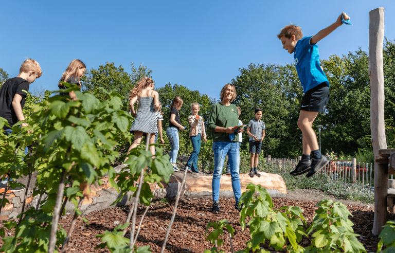 Kinderen spelen buiten in een natuurlijke omgeving met een volwassen begeleider onder een blauwe lucht.