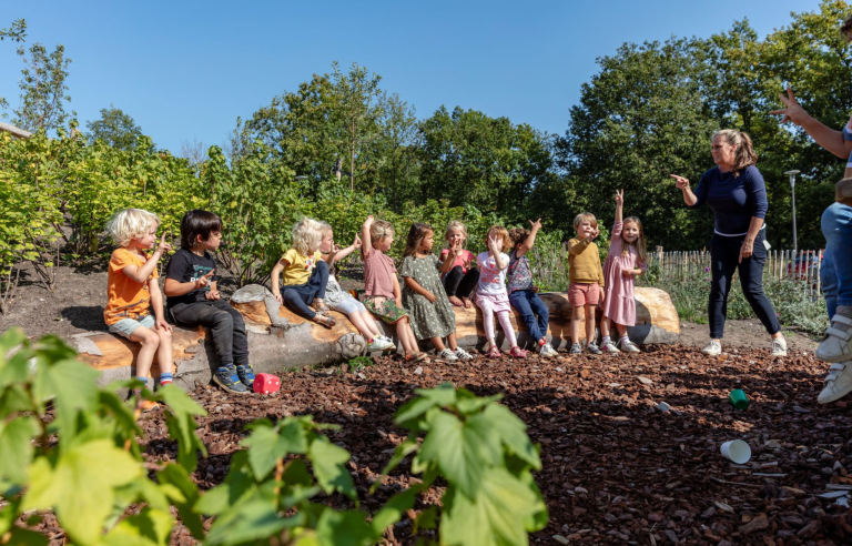 Kinderen zitten buiten op boomstammen met een volwassene; ze spelen een spel.