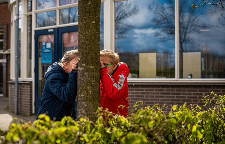 Twee kinderen spelen verstoppertje achter een boom voor een schoolgebouw.