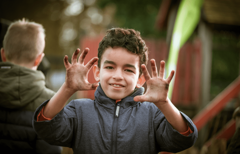 Een jongen met vieze handen glimlacht buiten, terwijl een andere jongen rugwaarts staat.