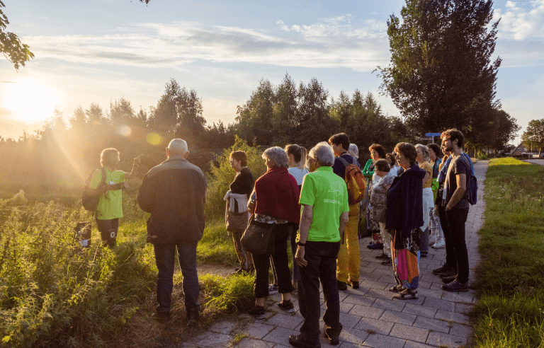 Groep mensen luistert naar een gids tijdens een zonsondergang in een park.