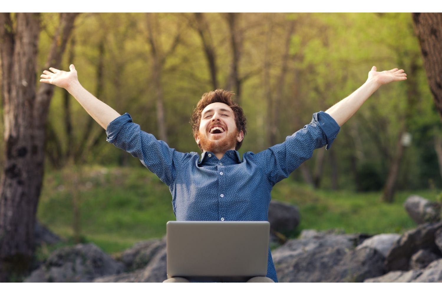 Man juicht met laptop op schoot in een groen bos.