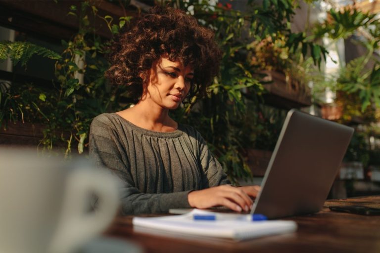 Vrouw werkt buiten op een laptop, omgeven door planten, notitieblok en pen op tafel.