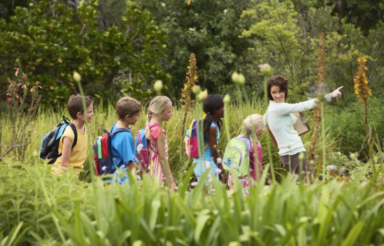 Groep kinderen met rugzakken volgt een vrouw, wijzend in een weelderige, groene omgeving.