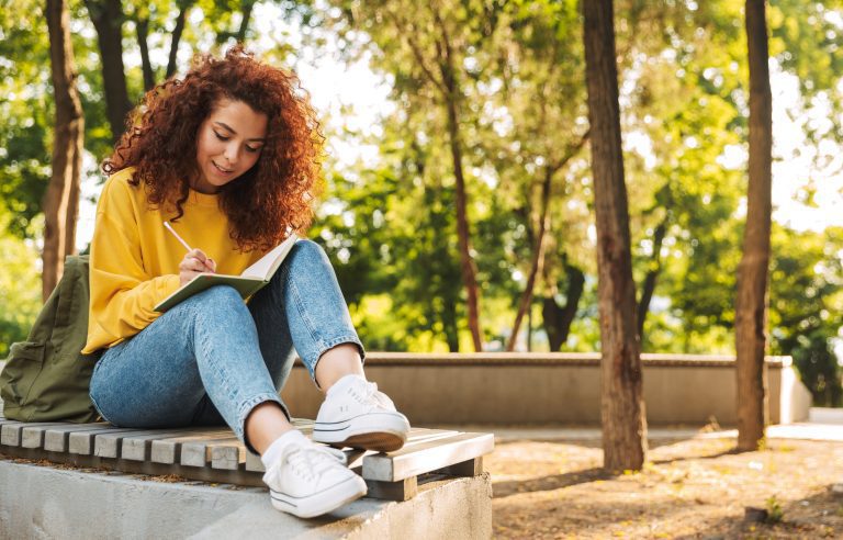 Vrouw schrijft in notitieboekje terwijl ze op een bank in een zonnig park zit.