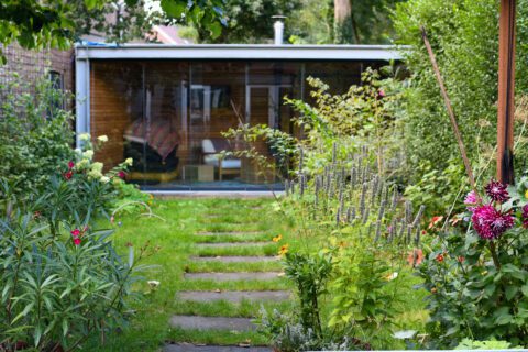 Pad door groene tuin naar een glazen schuurtje, omlijst door diverse planten en bloemen.