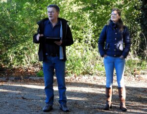 Twee mensen buiten, iemand spreekt terwijl de ander luistert, omringd door groene struiken.