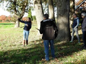 Persoon toont iets aan anderen bij bomen in een park, met herfstsfeer en een huis op de achtergrond.