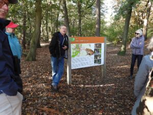 Een gids geeft uitleg bij een informatiebord in een bosrijke omgeving, omringd door bezoekers.