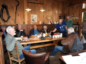 Groep oudere mannen aan tafel in houten kamer, drinken koffie en praten.