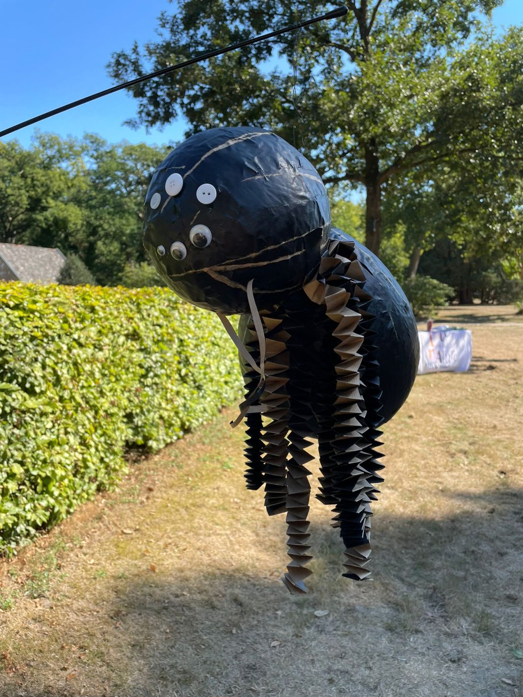 Een zwarte papieren spin met kartonnen poten, opgehangen in een zonnig park.