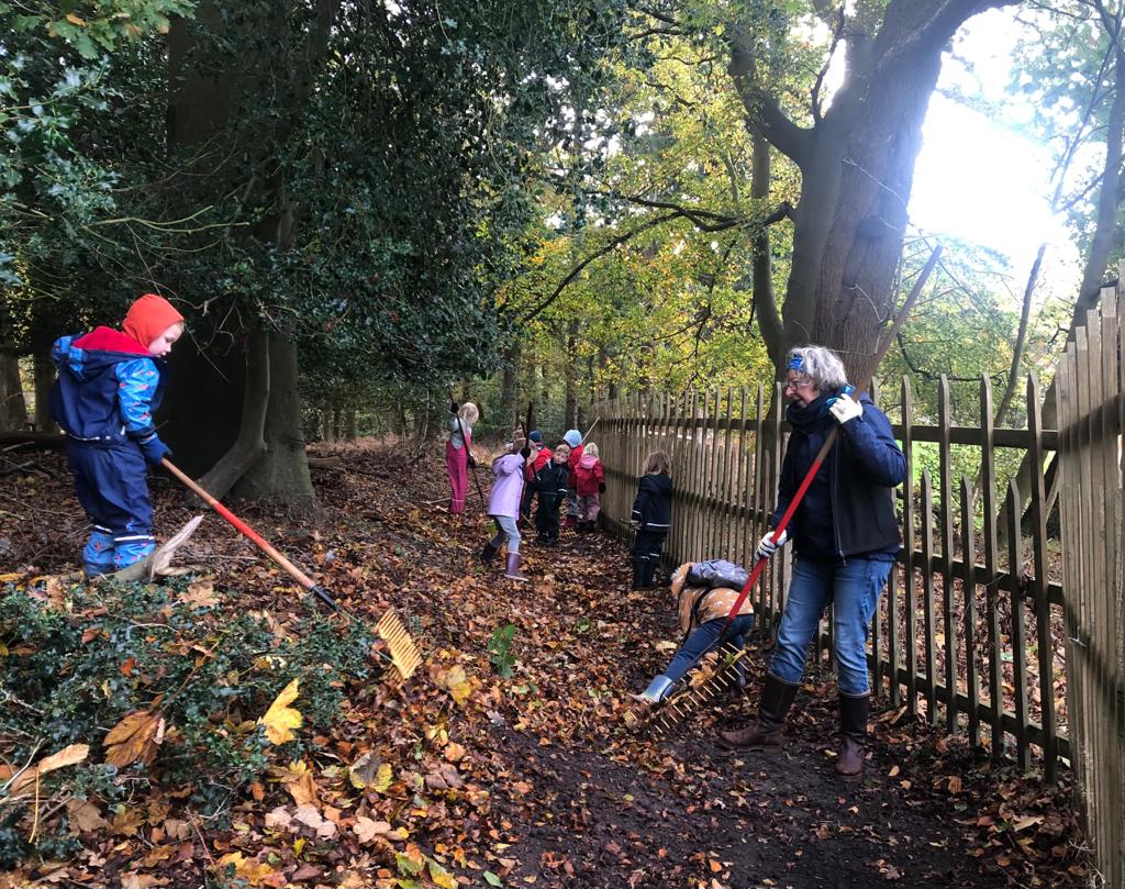 Kinderen harken bladeren op bospad, begeleid door een volwassene langs een hek.