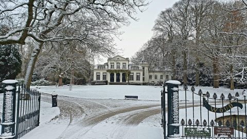 Omgracht landhuis in sneeuw, omgeven door bomen, met een ijzeren poort op de voorgrond.