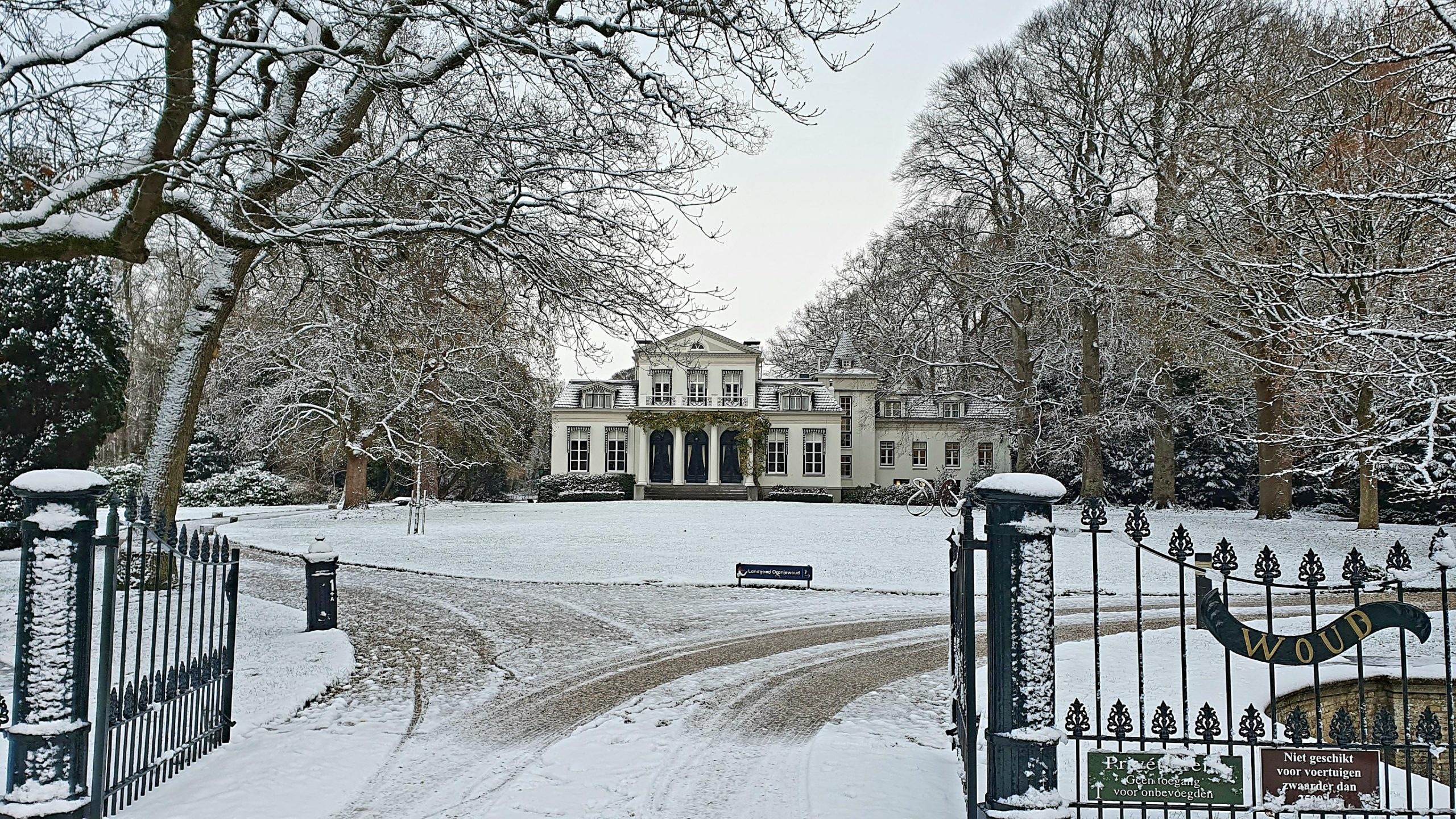 Omgracht landhuis in sneeuw, omgeven door bomen, met een ijzeren poort op de voorgrond.