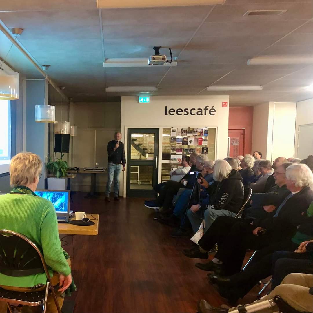 Presentatie in een leescafé; spreker voor een volle zaal met publiek, vrouw bedient laptop.