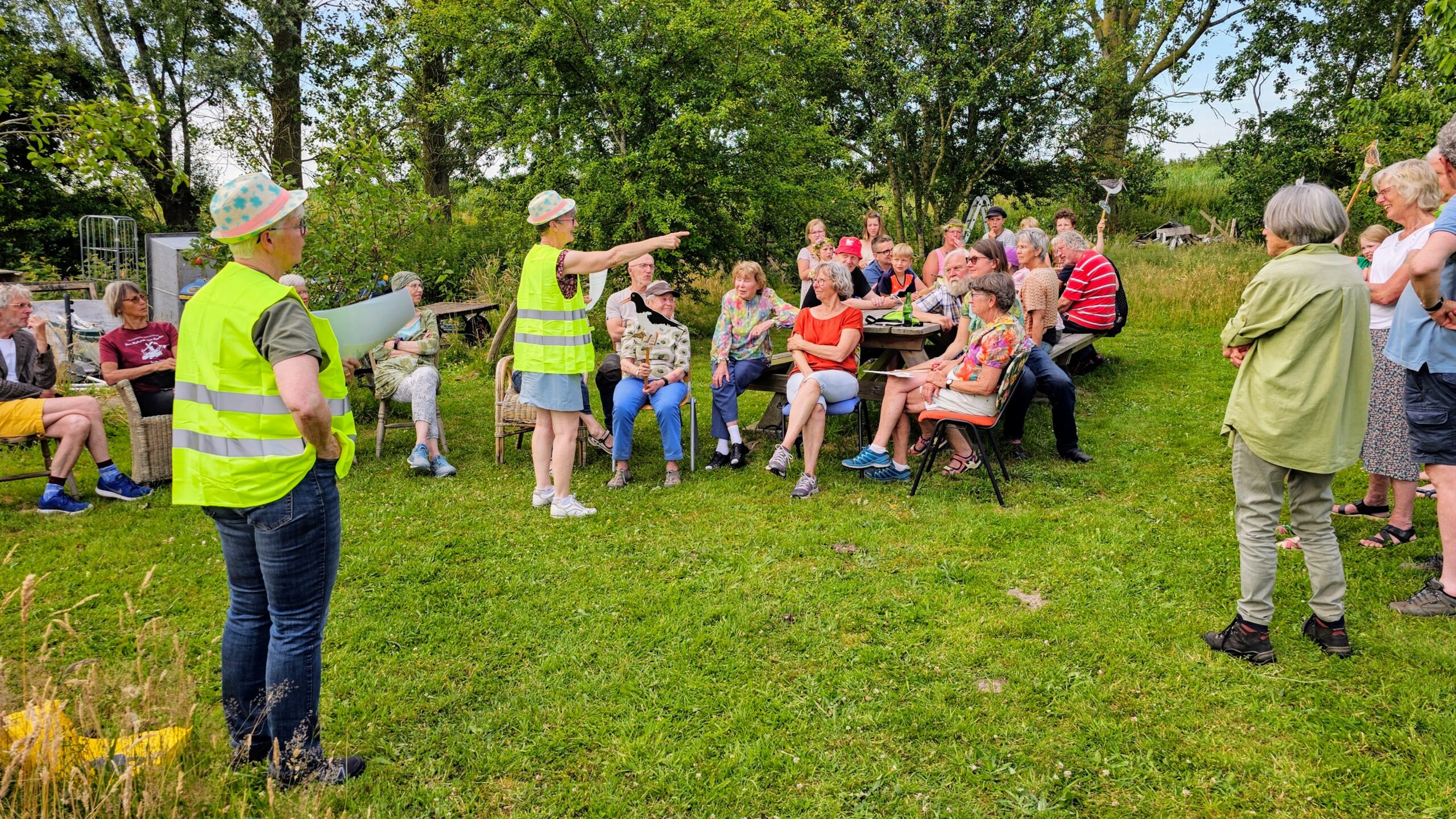 Een groep mensen luistert naar een spreker in gele hesjes buiten op een grasveld.