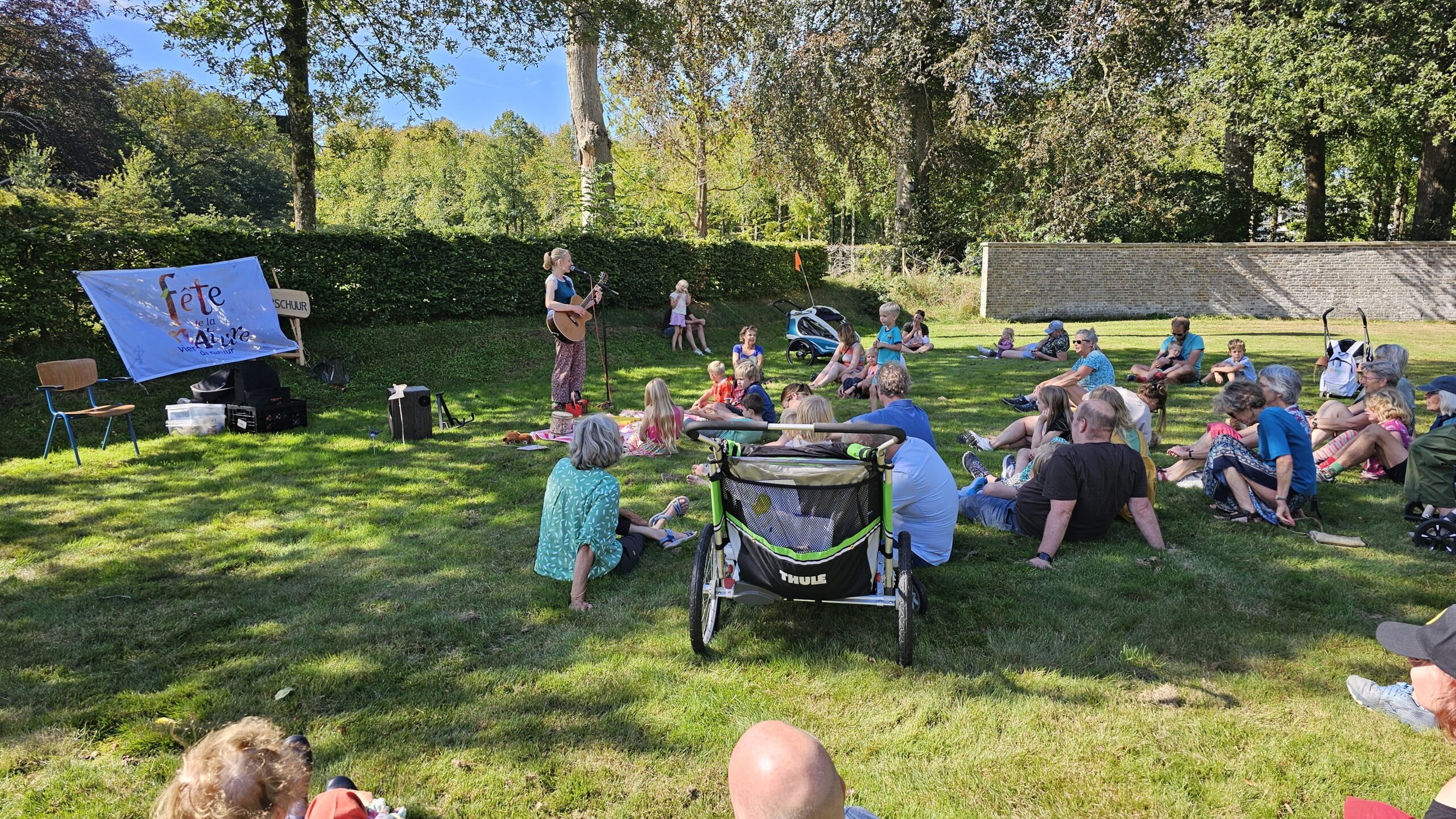 Mensen zitten buiten op gras, luisteren naar een muziekoptreden. Banner met "Fête de la Nature".