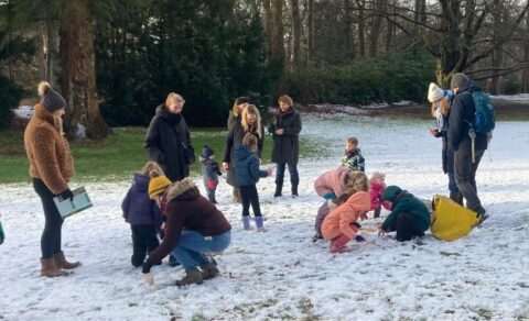 Groep volwassenen en kinderen speelt in de sneeuw in een park met bomen op de achtergrond.