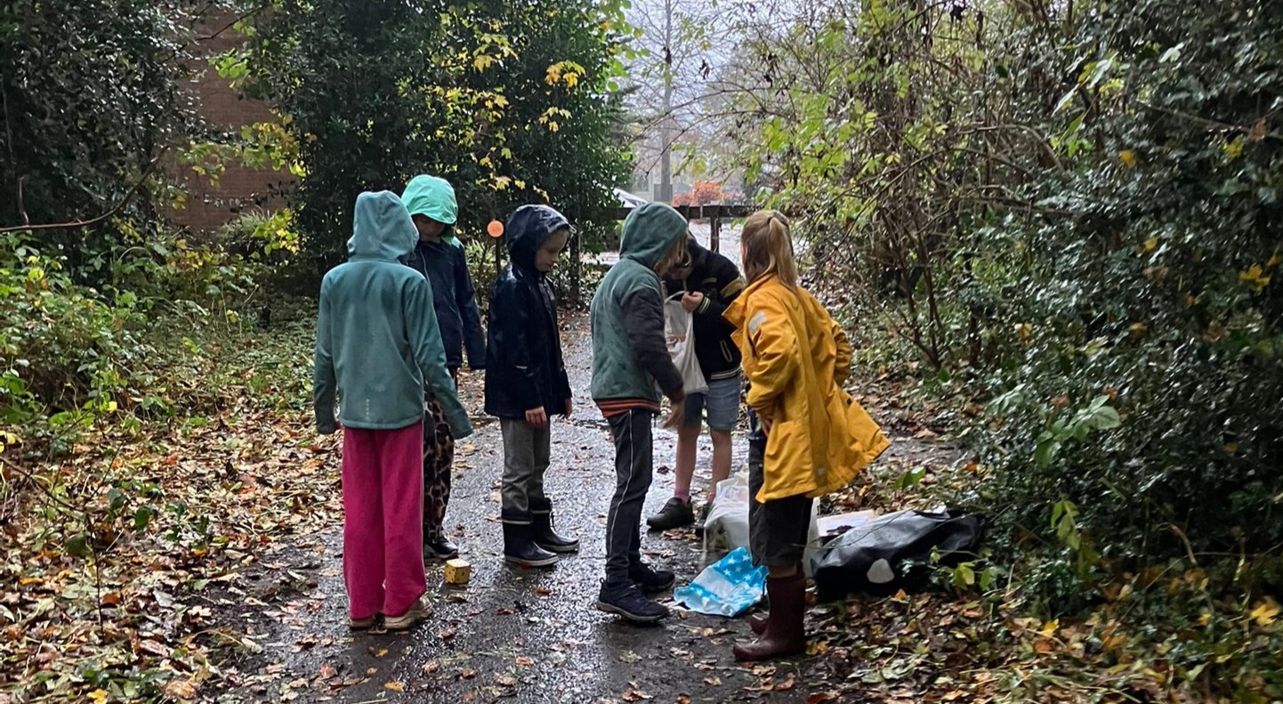 Kinderen in regenjassen verzamelen zich op een bosrijk pad, omgeven door herfstbladeren.