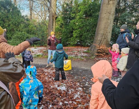 Kinderen en volwassenen spelen in een herfstig bos met bladeren en sneeuw op de grond.