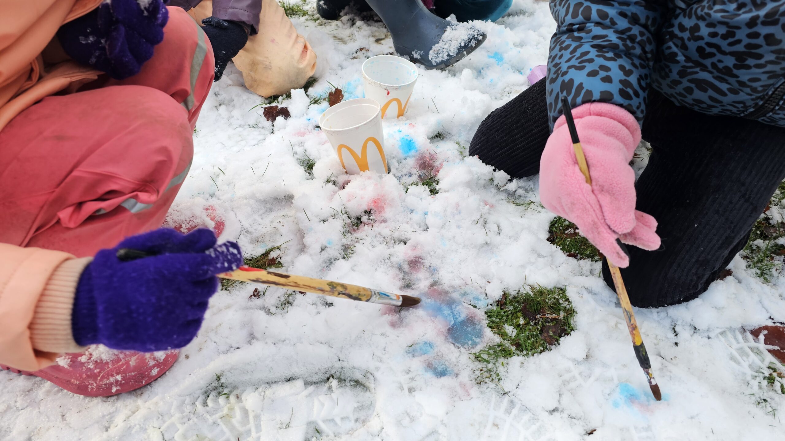 Kinderen schilderen met kwasten op besneeuwd gras, McDonald's bekers als waterpotjes.