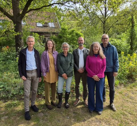 Een groep van zes mensen poseert voor een huis in een groene, bosrijke omgeving.