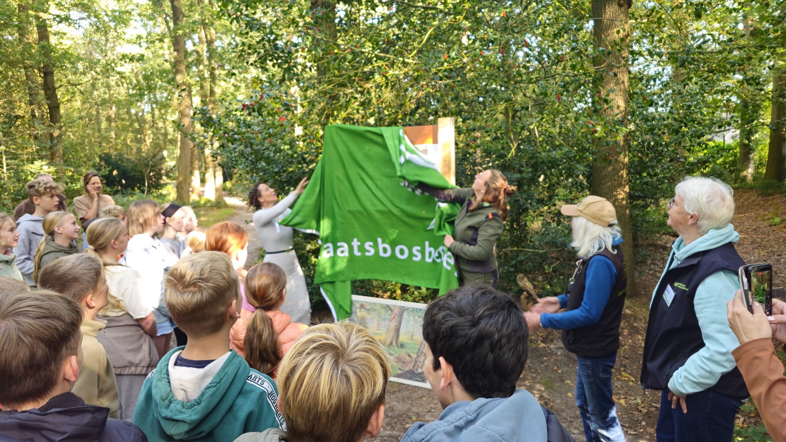 Groep mensen onthult bord in bos, kinderen kijken toe, groene doek met tekst "staatsbosbeheer".