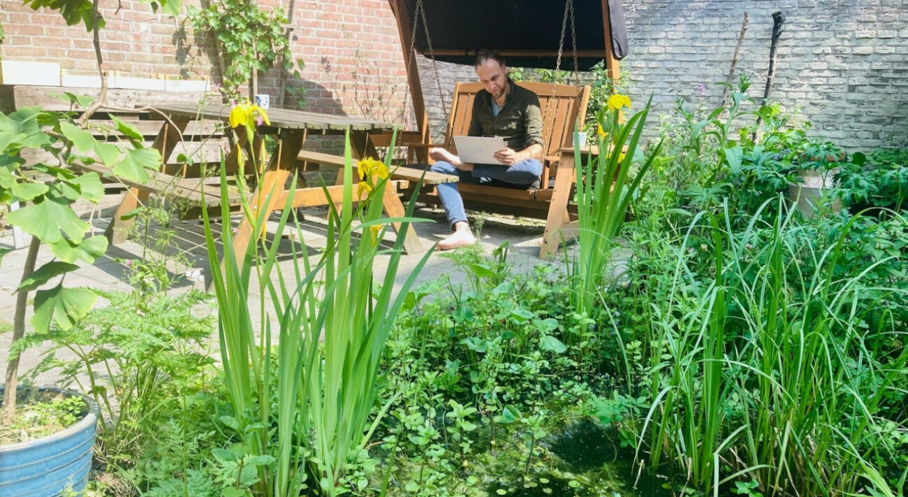 Man leest op een schommelbank in een groene, zonnige tuin met een vijver en houten tafel.