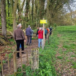 Groep wandelaars op een bosrijk pad naast een grasveld, bij een houten hek met gele bord.