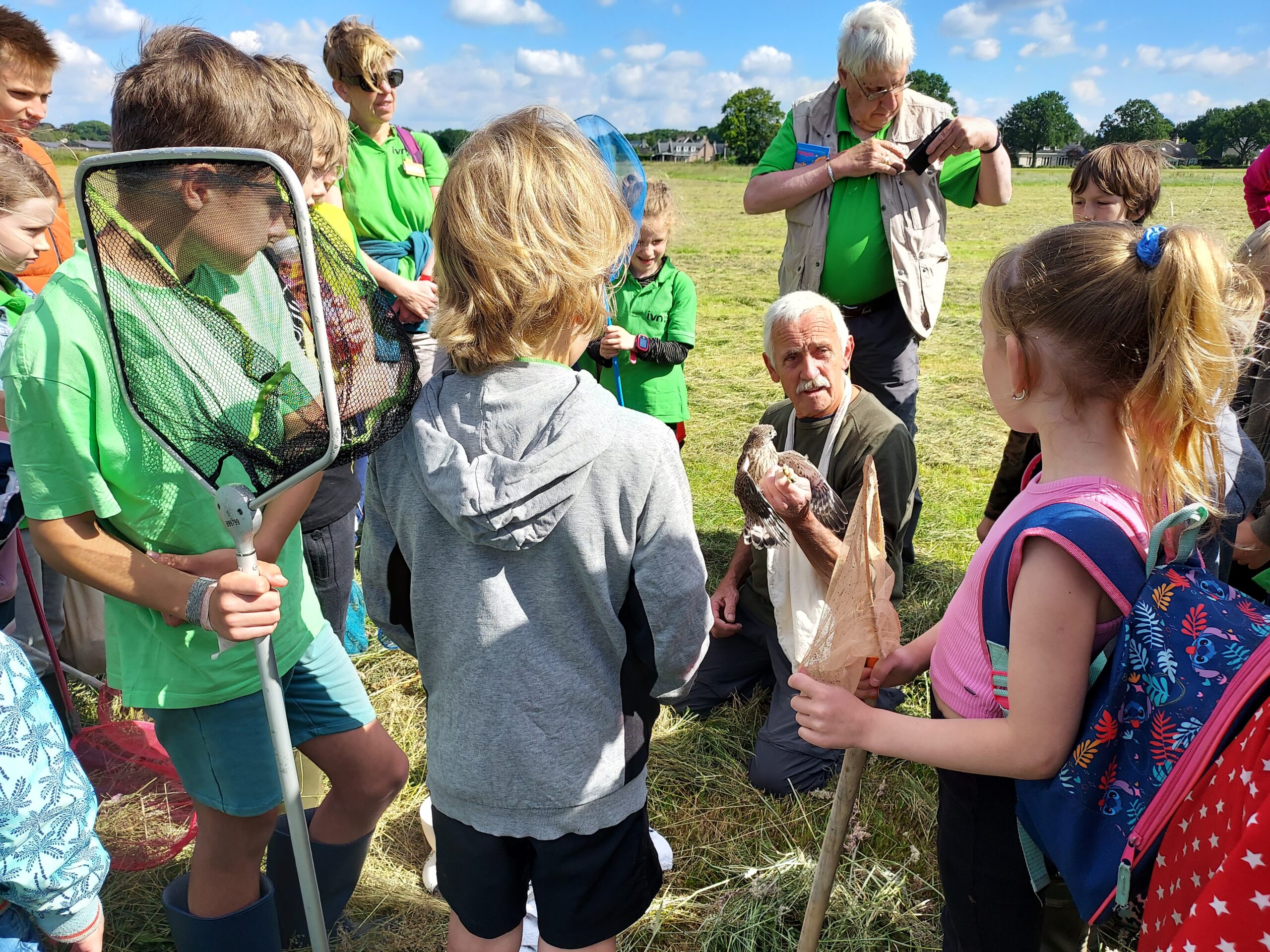 Groep kinderen observeert een man met een vogel op een grasveld onder een blauwe lucht.