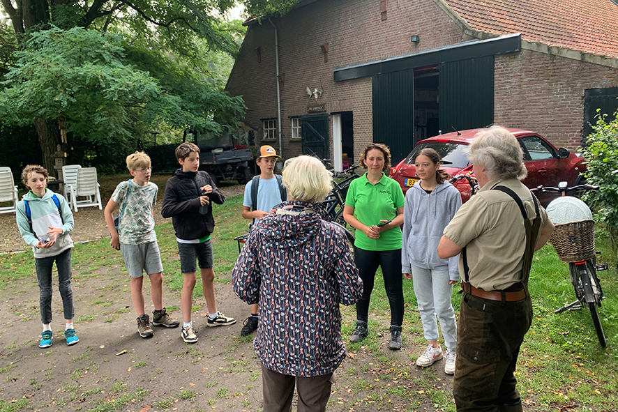 Groep mensen luistert naar een man bij een boerderij, met fietsen en een schuur op de achtergrond.