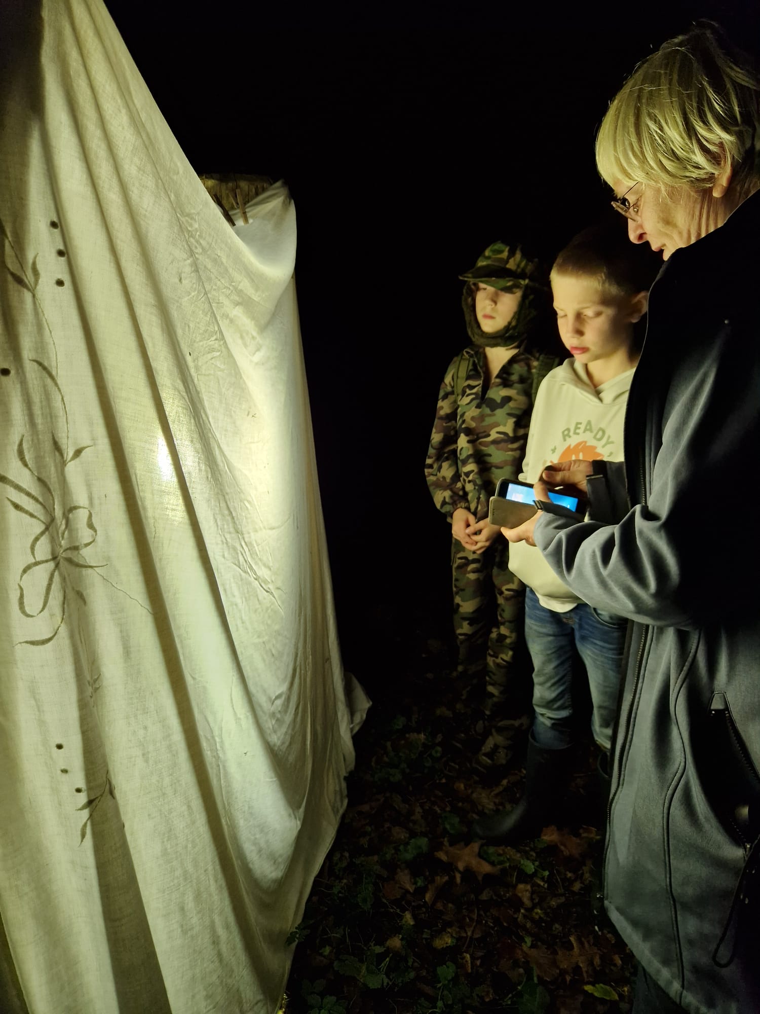 Drie mensen staan 's nachts buiten bij een verlicht laken met een patroon.