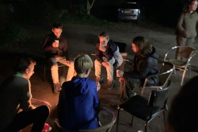 Groep mensen zit rond een kampvuur in het donker, met bekers in de hand.
