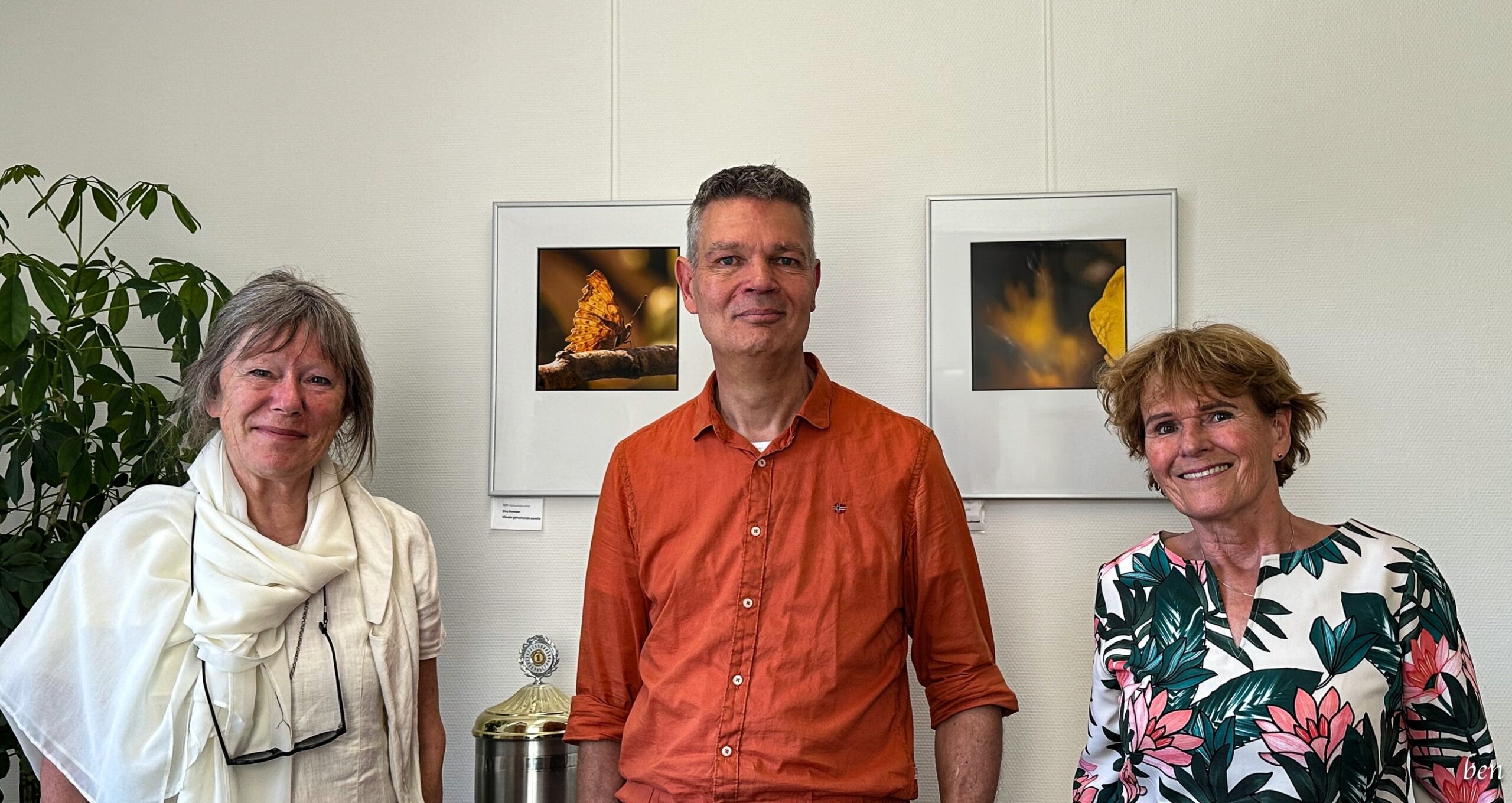 Drie mensen poseren voor kunstfoto's van vlinders aan de muur, met planten op de achtergrond.