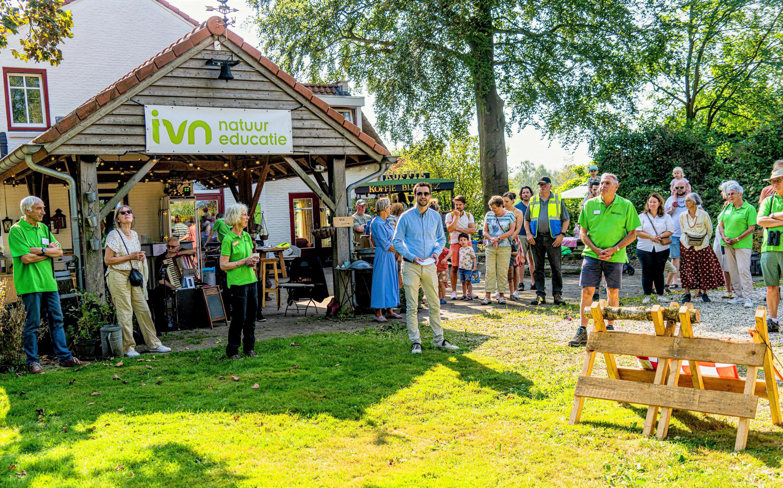 Bijeenkomst bij IVN-centrum met mensen en een spreker buiten in de zon, groen en houtomgeven.
