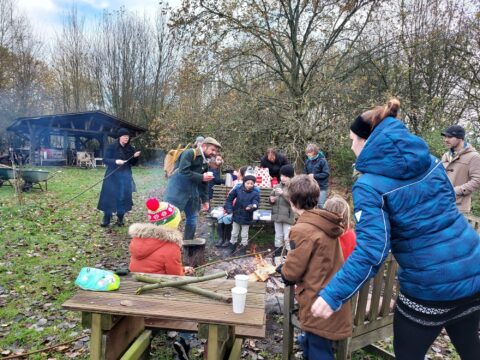 Groep mensen, waaronder kinderen, bij een kampvuur in een bosrijke omgeving.