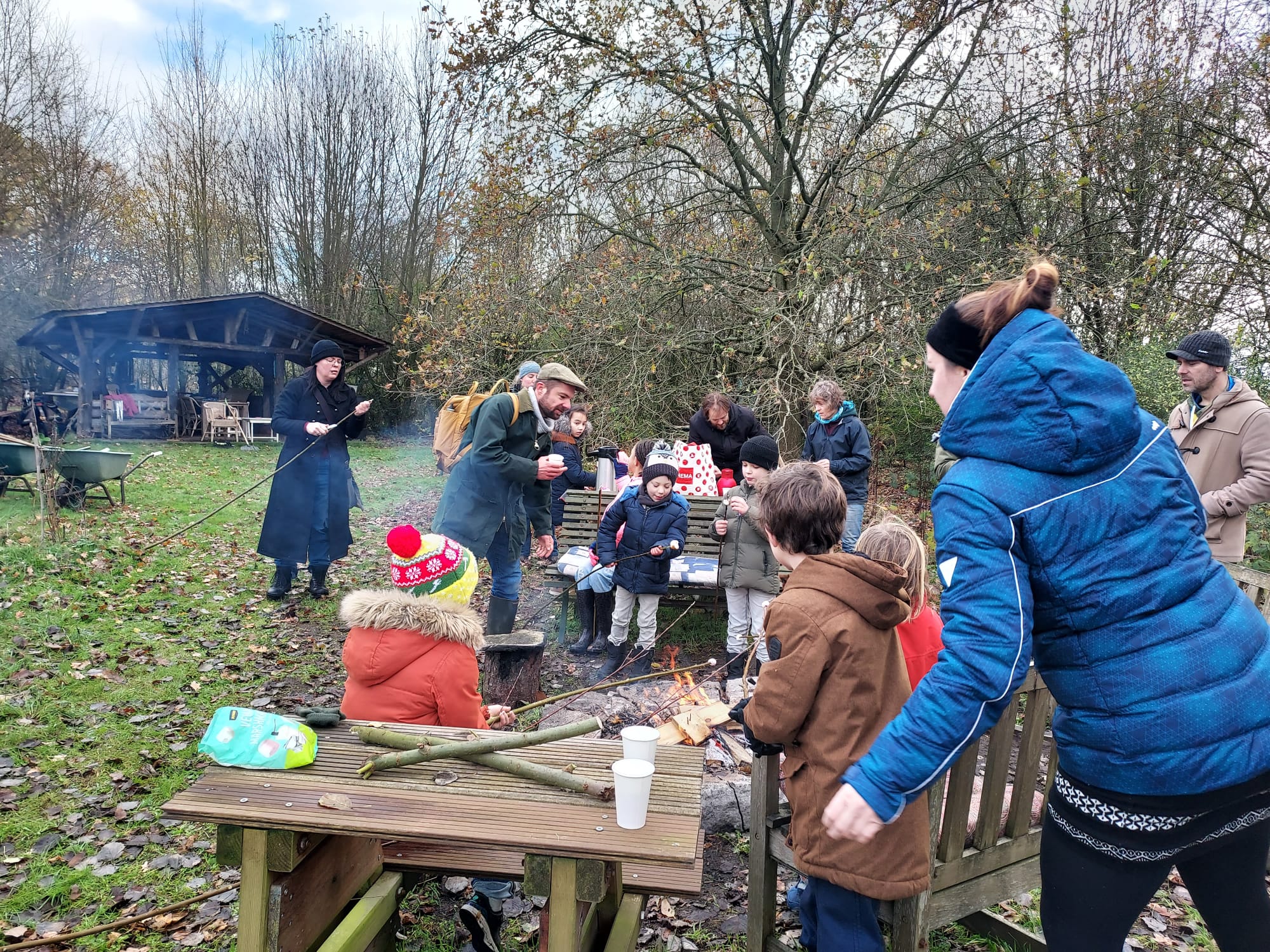 Groep mensen, waaronder kinderen, bij een kampvuur in een bosrijke omgeving.