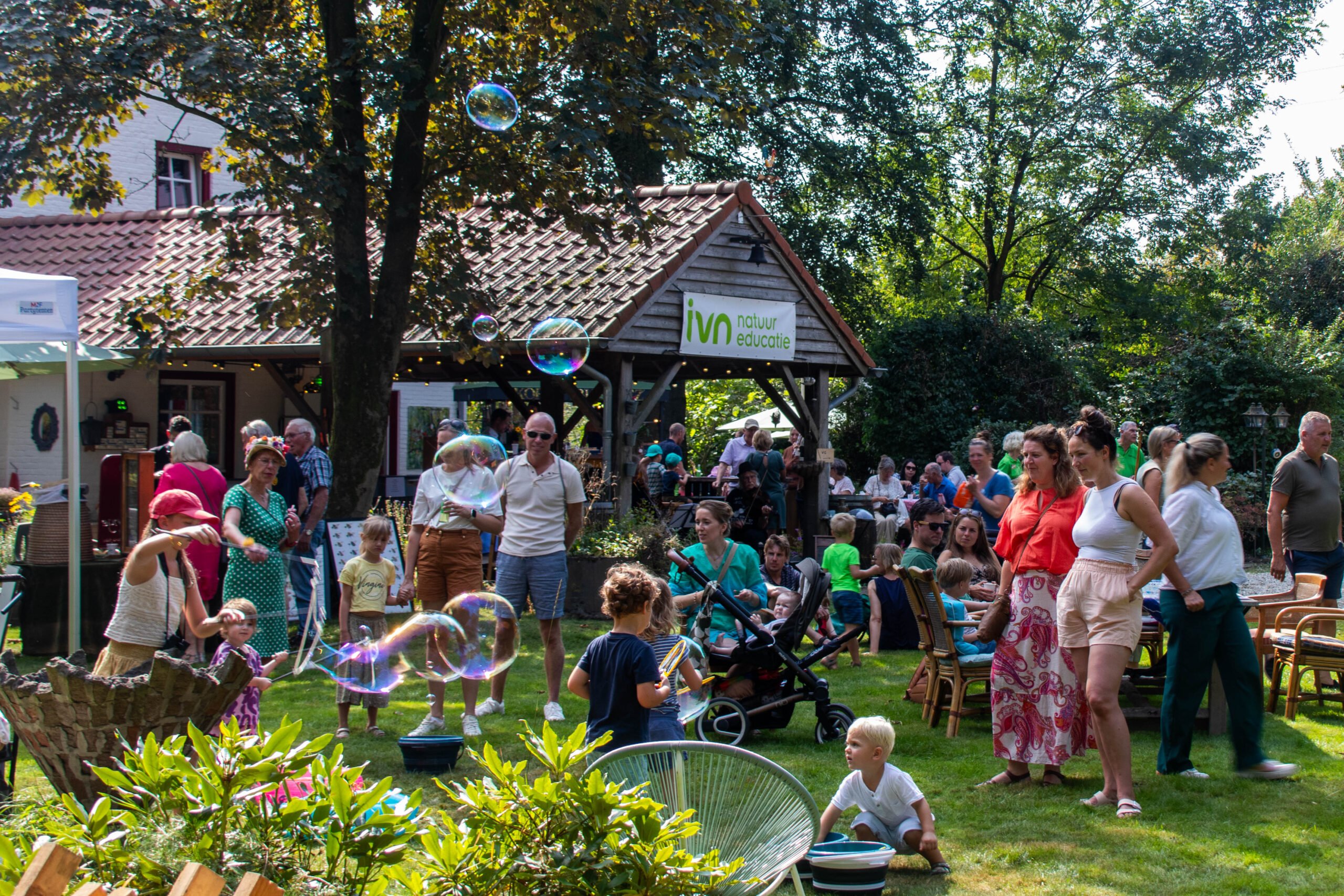 Een drukke tuin met mensen en kinderen die spelen en bellen blazen tijdens een evenement bij IVN Natuureducatie.