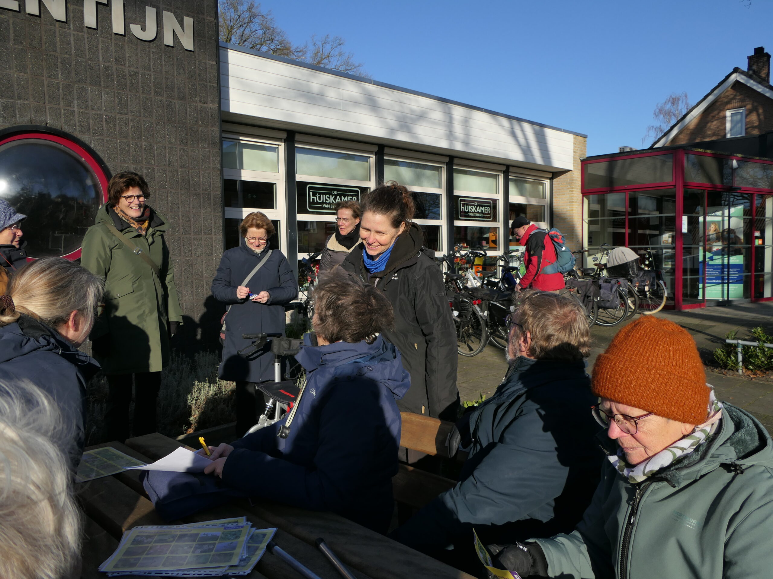 Groep mensen verzameld bij een gebouw, enkele met papieren, fietsen op de achtergrond.