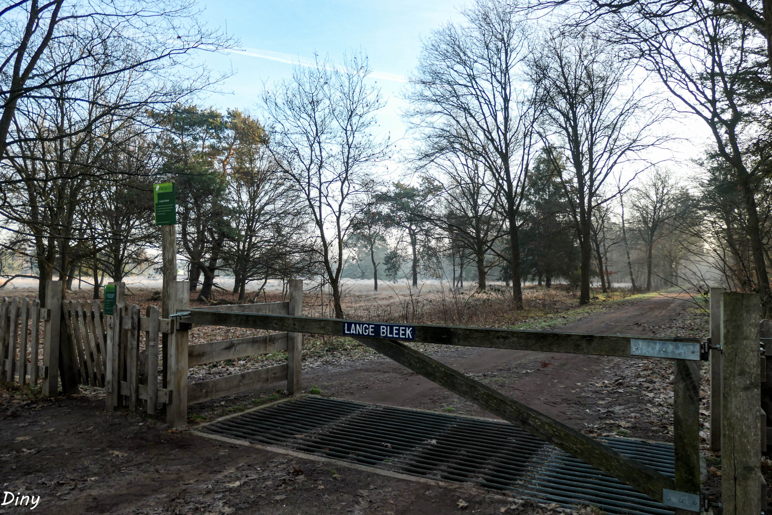 Houten poort met bord "Lange Bleek", plattelandsweg, bomen en open veld onder een heldere lucht.