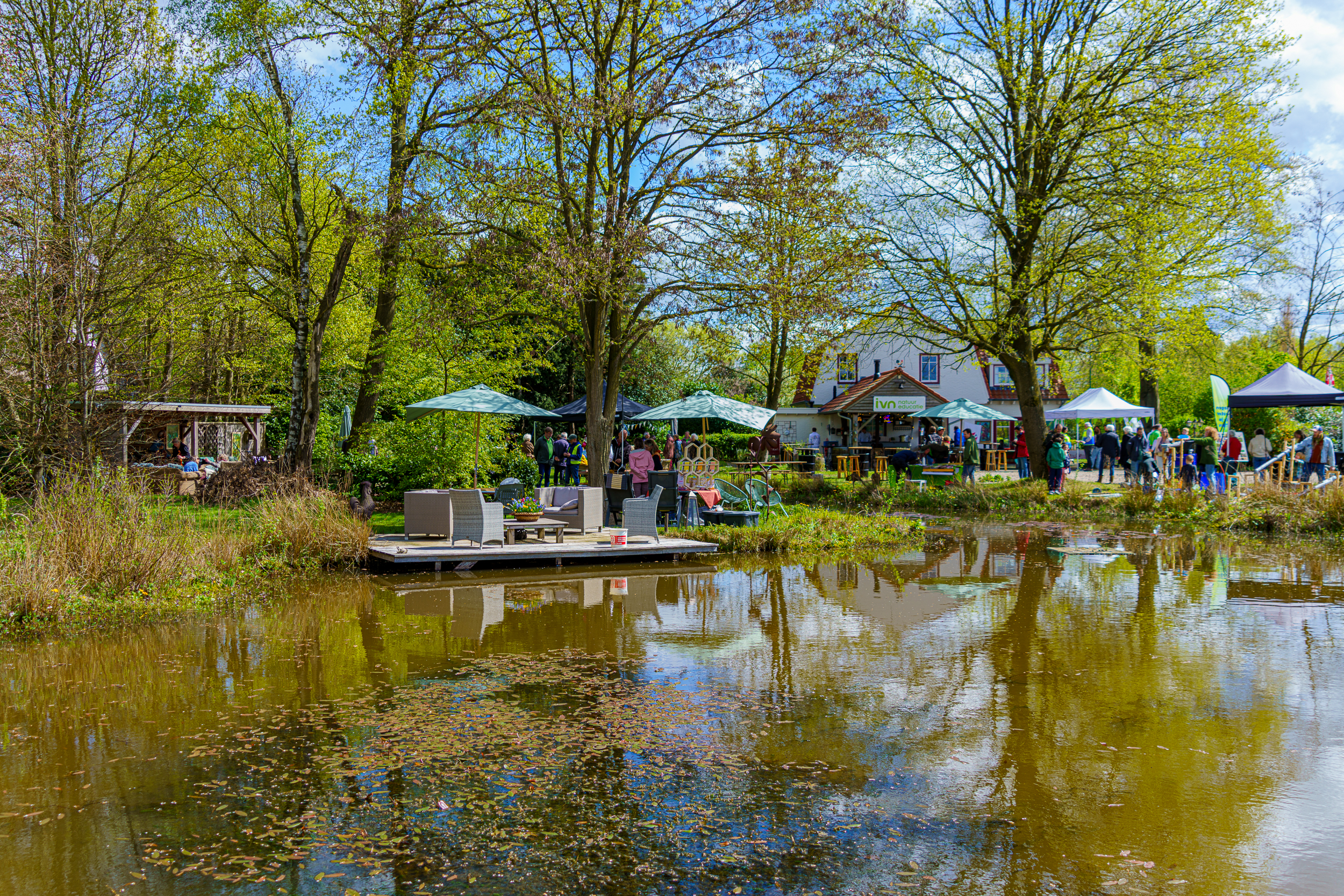 Tuinfeest bij een vijver met groene bomen, stoelen, parasols en mensen bij kraampjes.