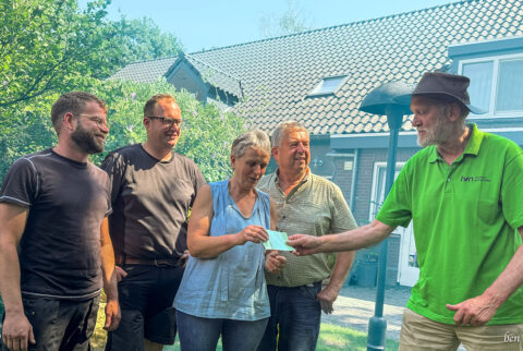 Groep mensen, buiten bij een huis, een persoon overhandigt iets aan een vrouw.