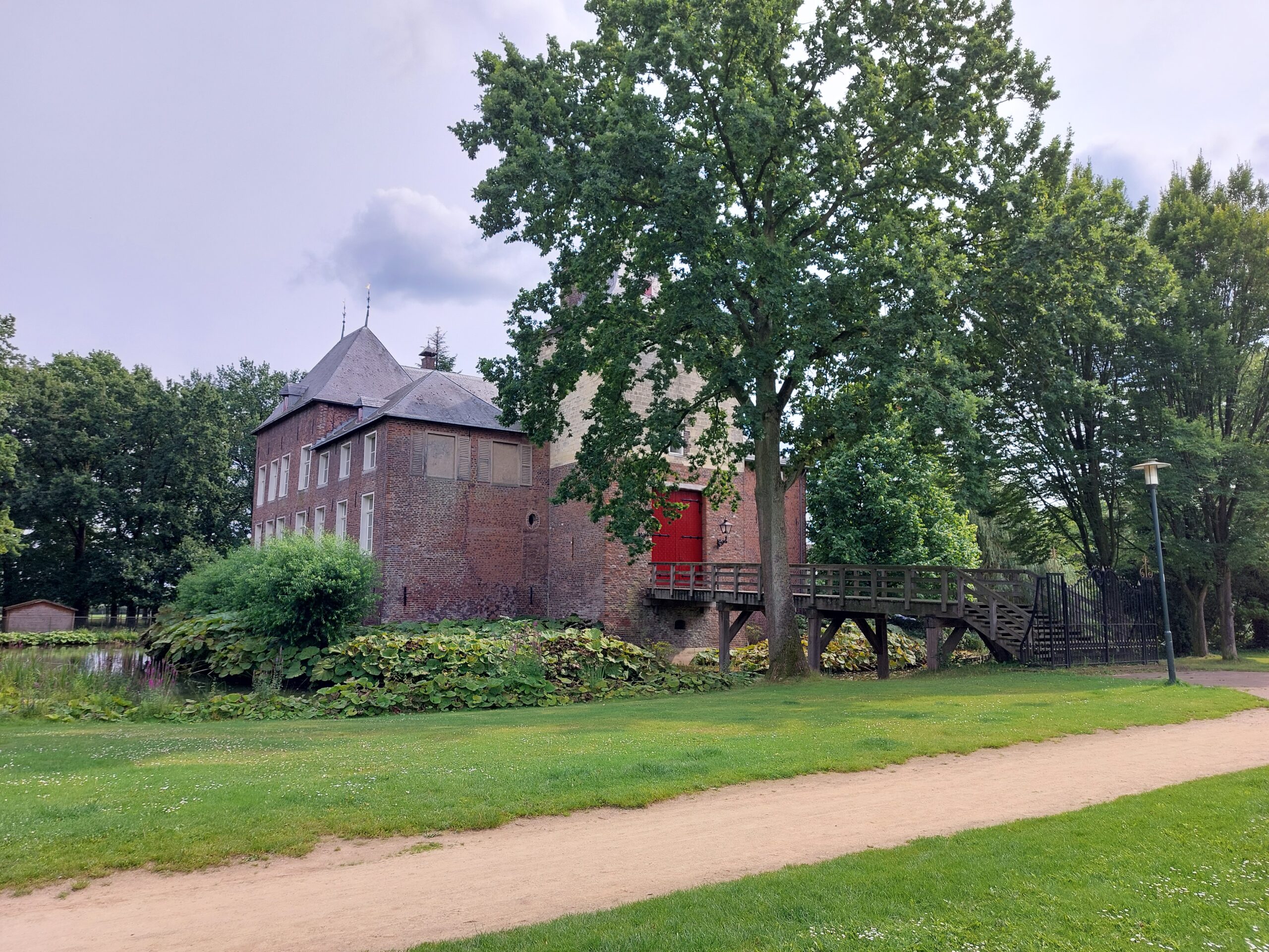 Bakstenen gebouw met rode deur, houten loopbrug, omgeven door bomen en een grasveld.