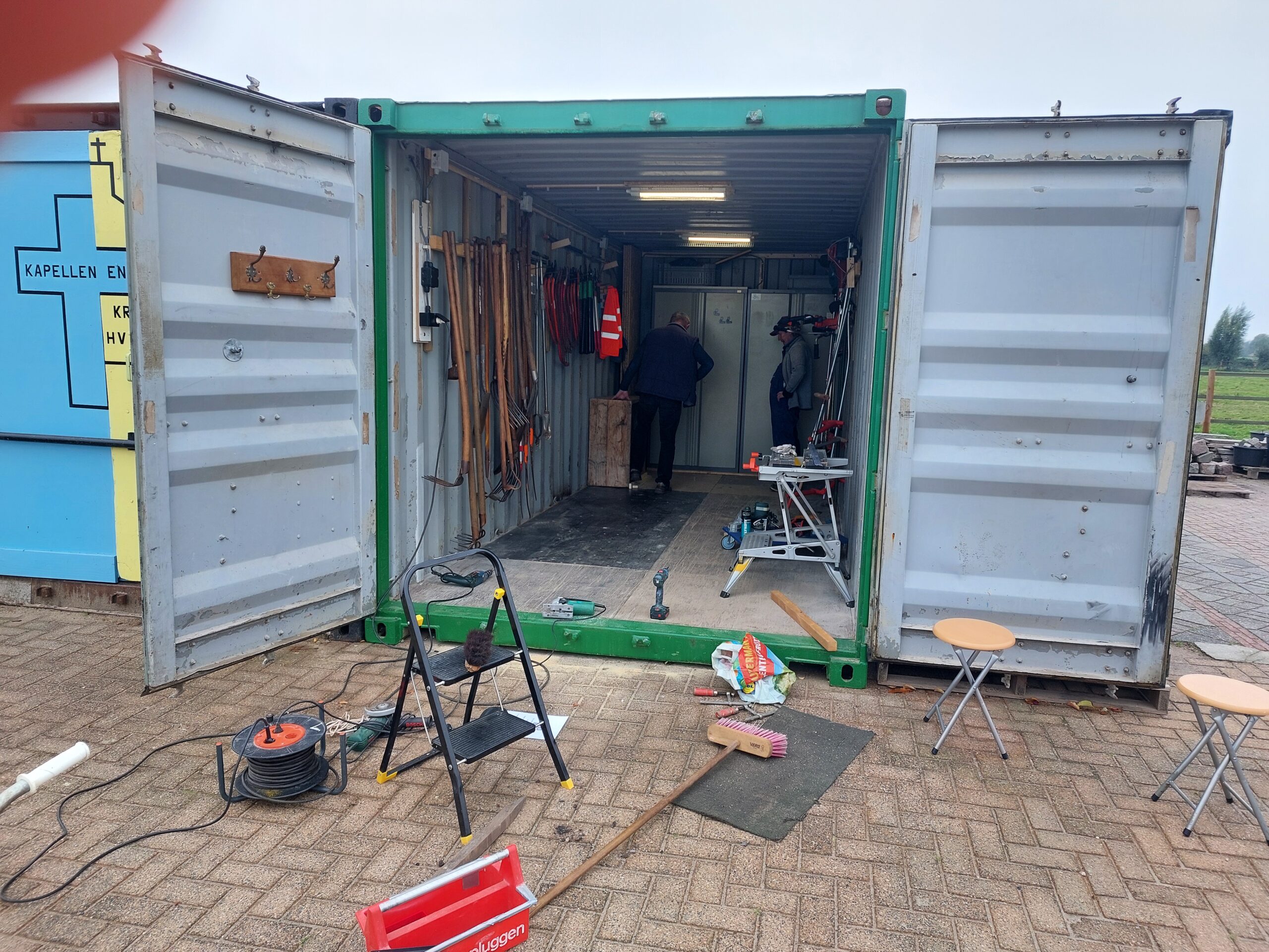 Een open container met gereedschap, twee mensen binnenin, en diverse spullen buiten op een stoep.