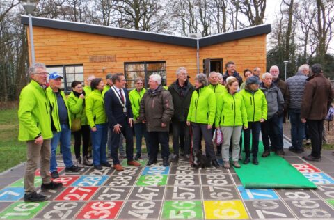 Groep mensen voor houten natuurhut, sommigen dragen felgroene jassen, op een kleurrijk genummerd pad.